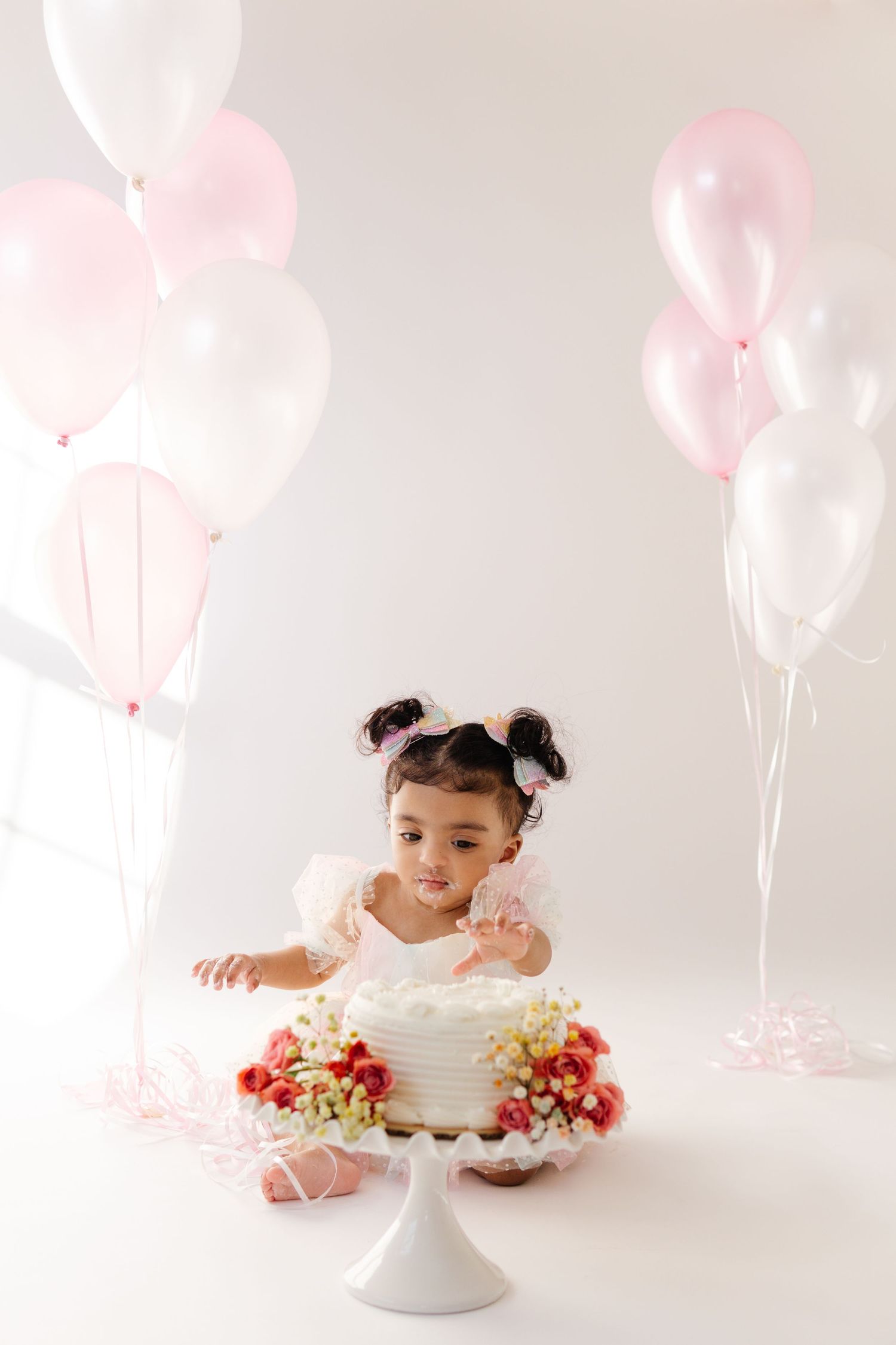 A toddler in a tulle dress reaches for a white birthday cake surrounded by pink balloons in a bright studio.
