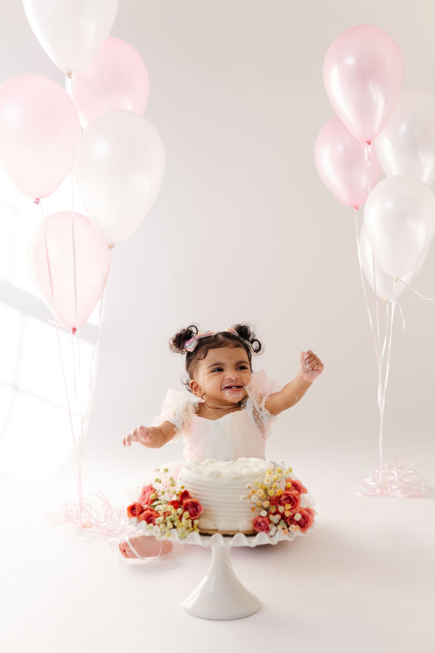 A small toddler in white dress sits happily next to a white cake decorated with flowers, surrounded by pink and white balloons.