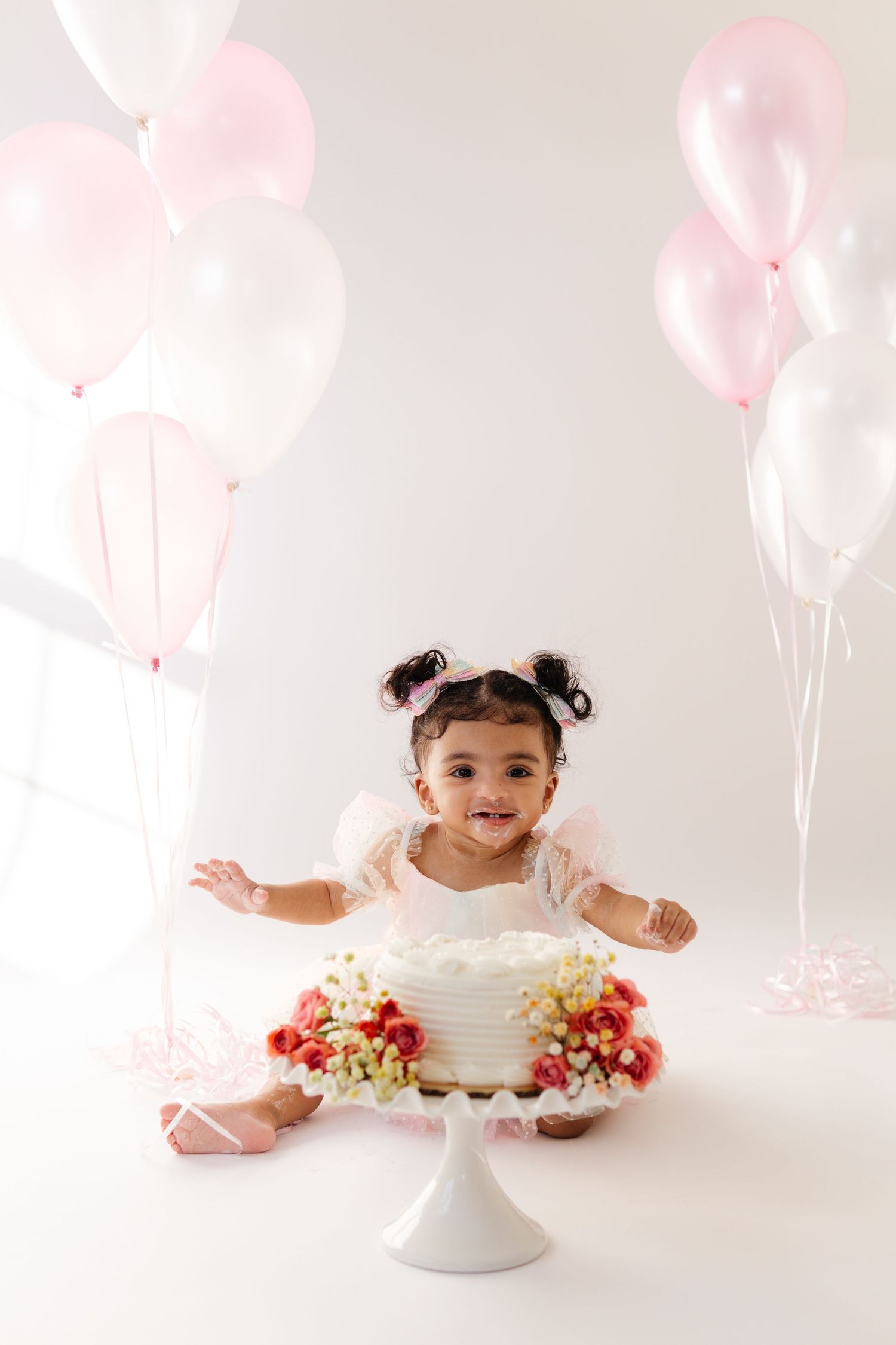 Sweet cake smash photo setup with white cake on pedestal, pink balloons, and white backdrop for special celebration.