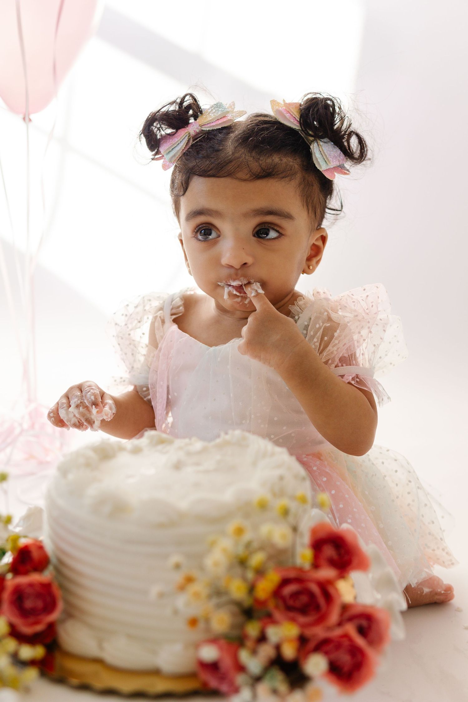 Baby in pink dress eating birthday cake surrounded by roses and party decorations at cake smash in studio session in Cherry Hill, NJ.
