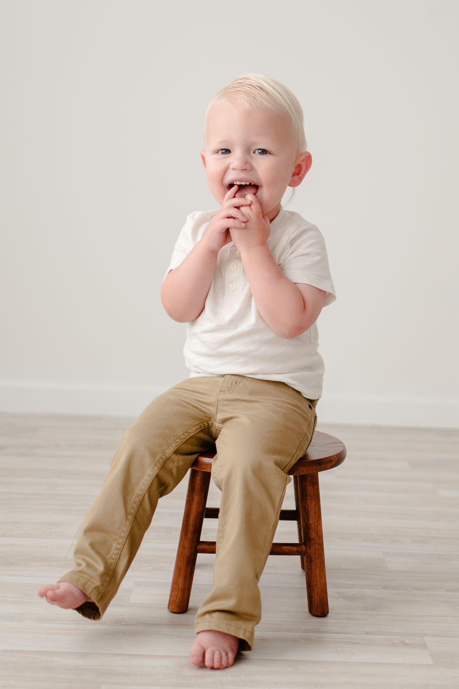 A toddler in a white t-shirt and khaki pants sits on a wooden stool against a neutral background, barefoot and giggling.