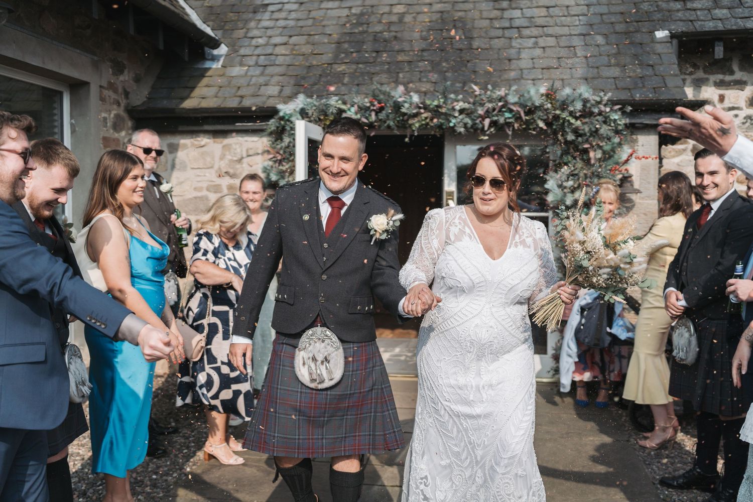 Wedding guests celebrate with the newlyweds outside a stone building, with the groom wearing a traditional Scottish kilt.