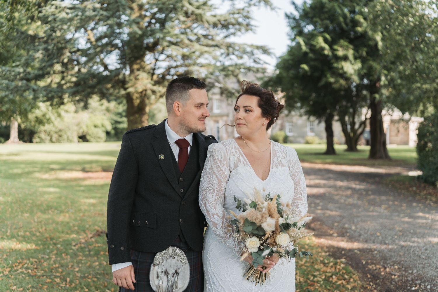 A couple shares a romantic moment in a park setting, with the bride holding a beautiful neutral-toned floral bouquet.