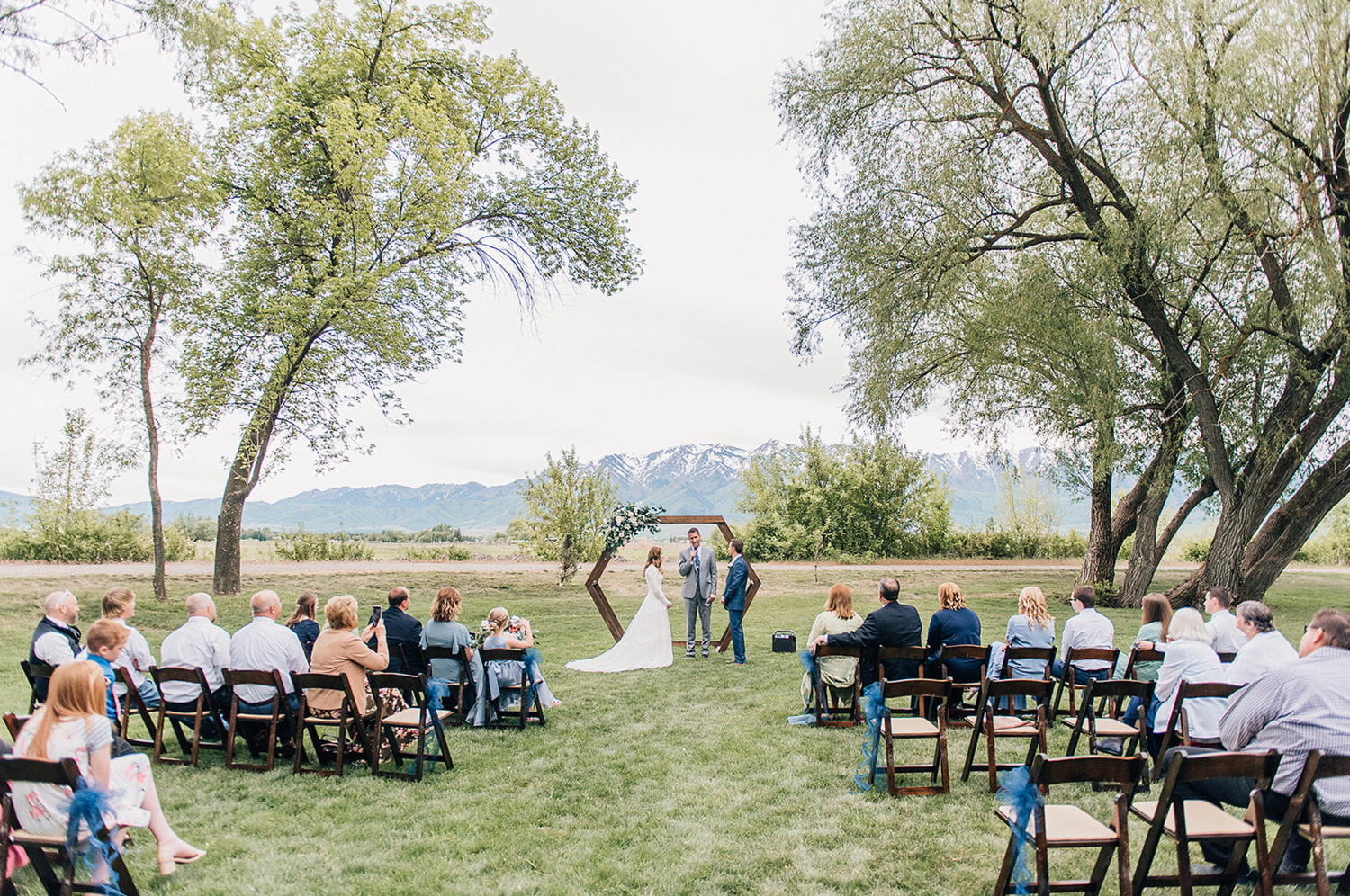 A scenic outdoor wedding ceremony setup with chairs on grass under willow trees with mountains in the background.