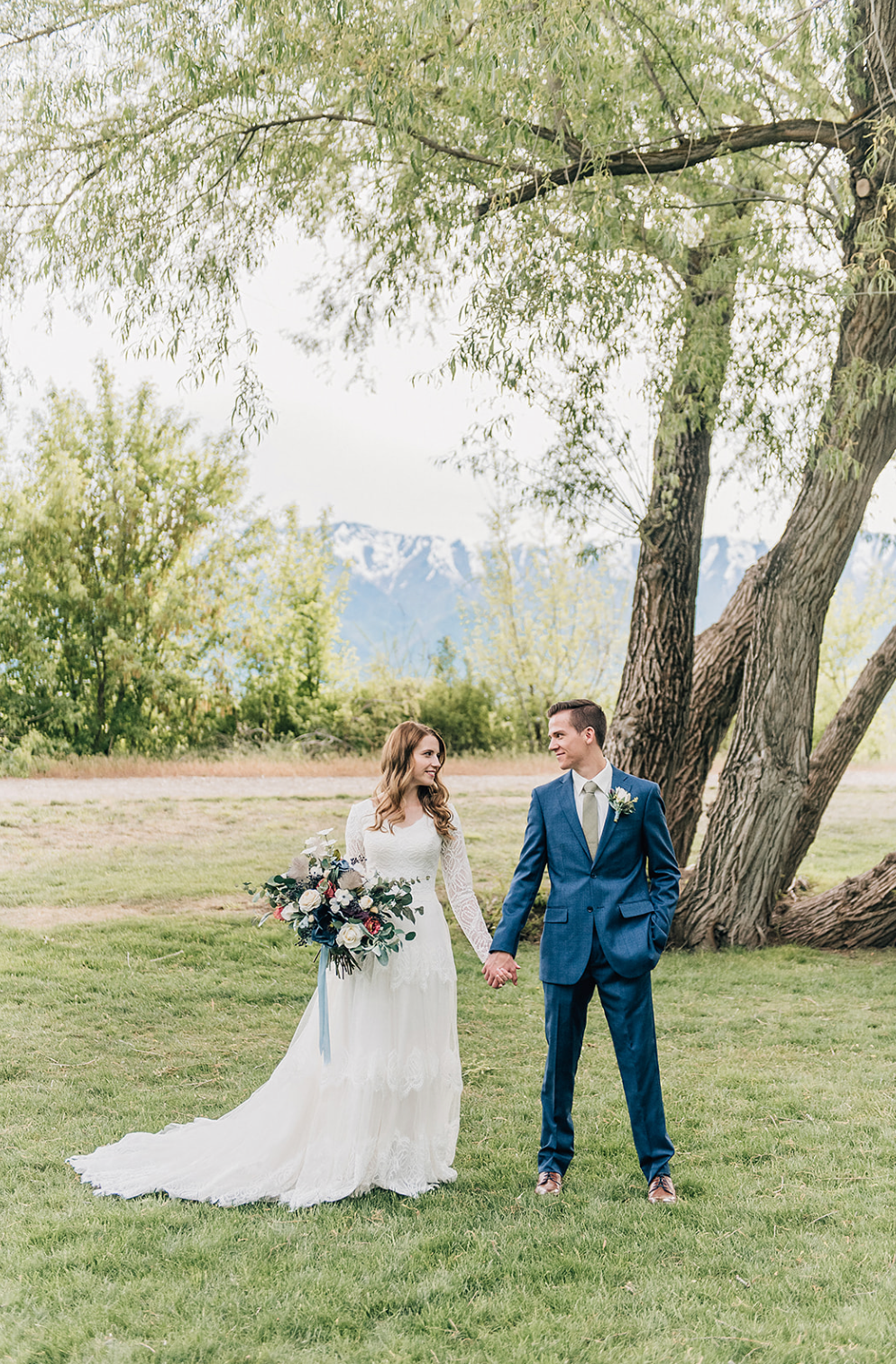 An elegant outdoor wedding photo against mountain views with two figures standing beneath a willow tree.