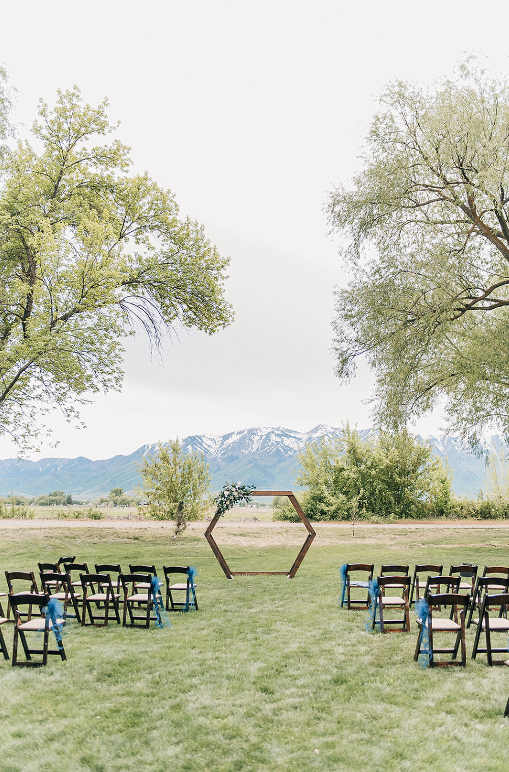 Outdoor wedding ceremony setup with hexagonal arch and blue chairs facing snow-capped mountains against green landscape.
