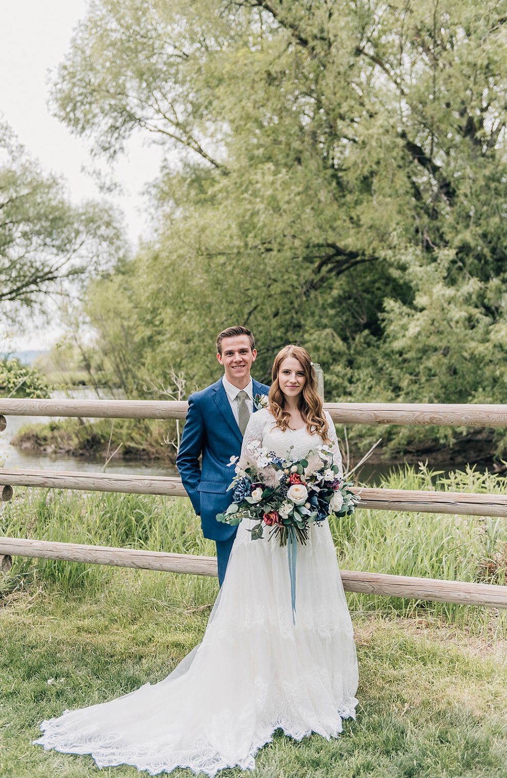A bride and groom pose together in a rustic outdoor setting with white lace dress, blue suit and natural greenery backdrop.