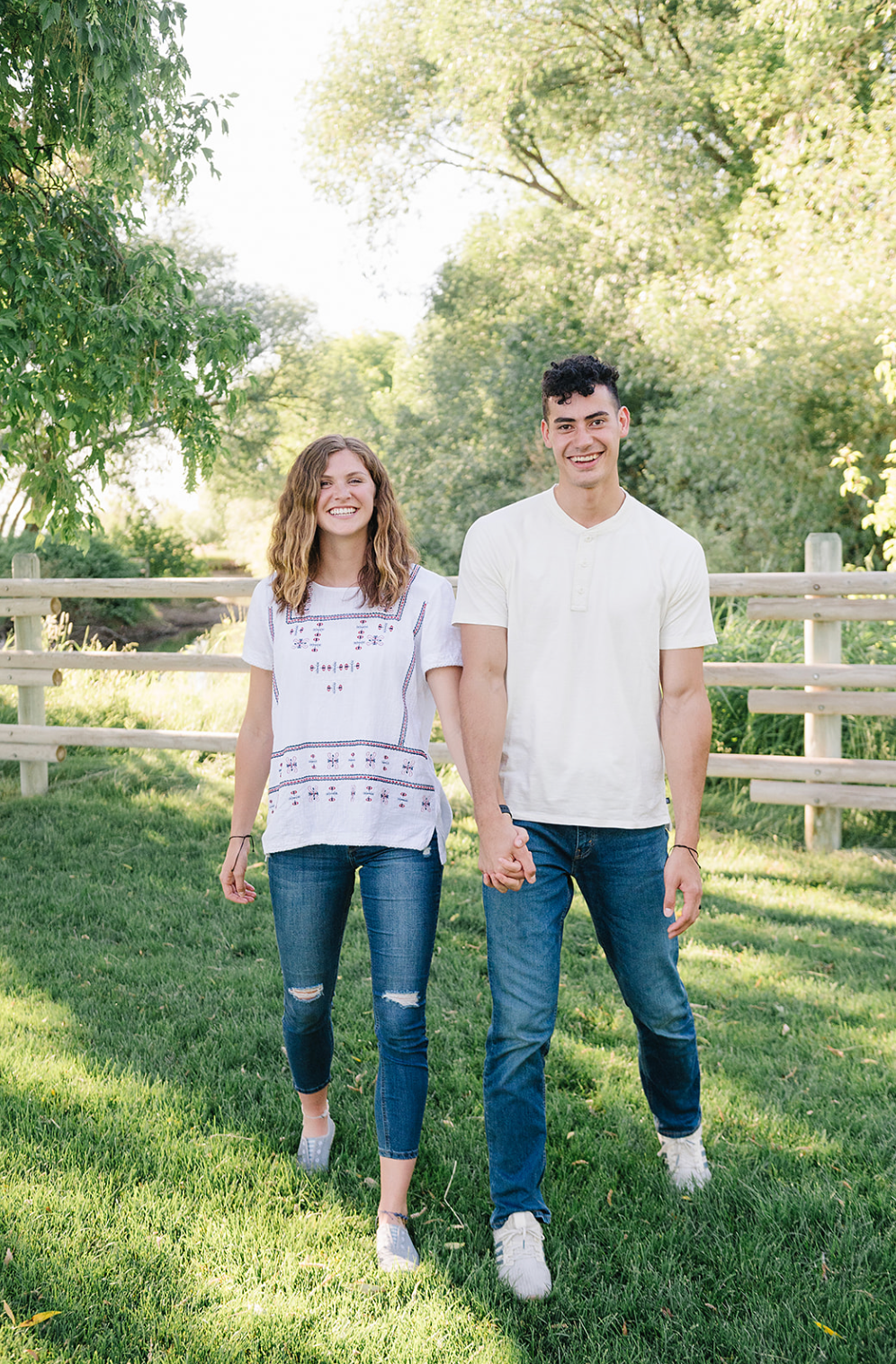 A couple walks hand in hand on green grass with a wooden fence and trees in the background.