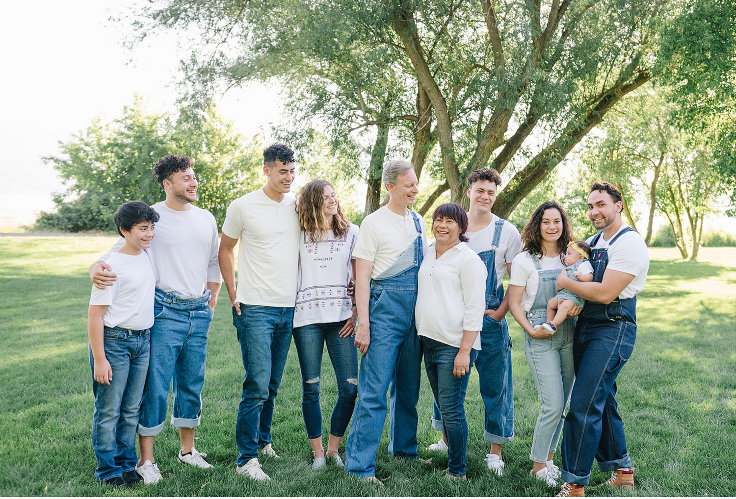 A group of people wearing coordinated white tops and blue jeans stand together on a grassy lawn under shady trees.