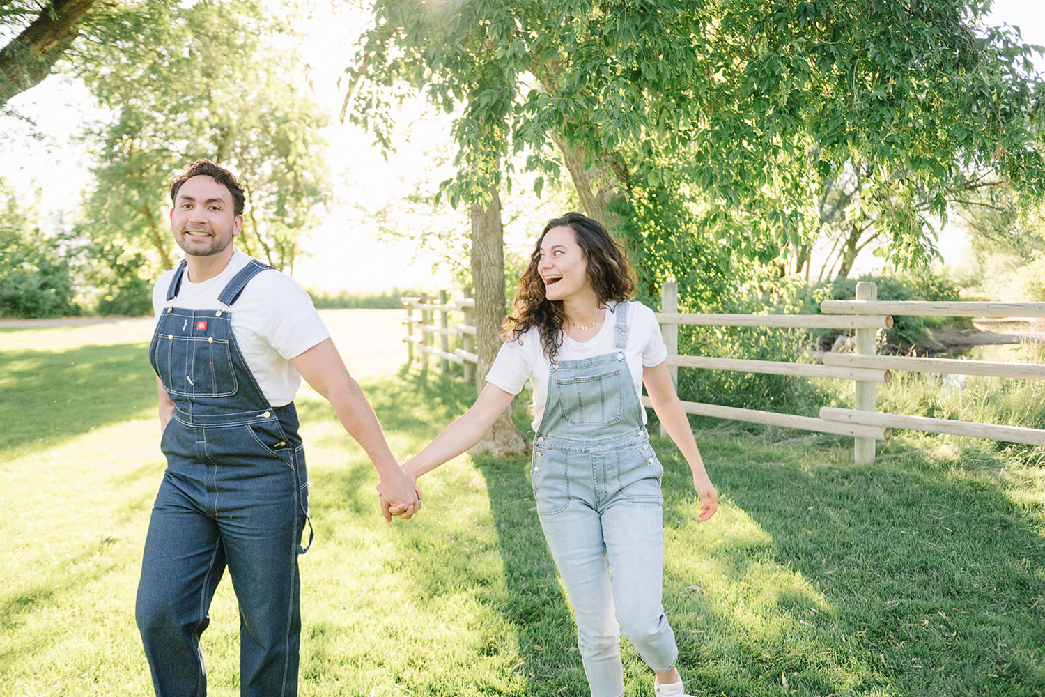 A couple wearing matching denim overalls walk hand in hand through a sunlit park on a bright summer day.