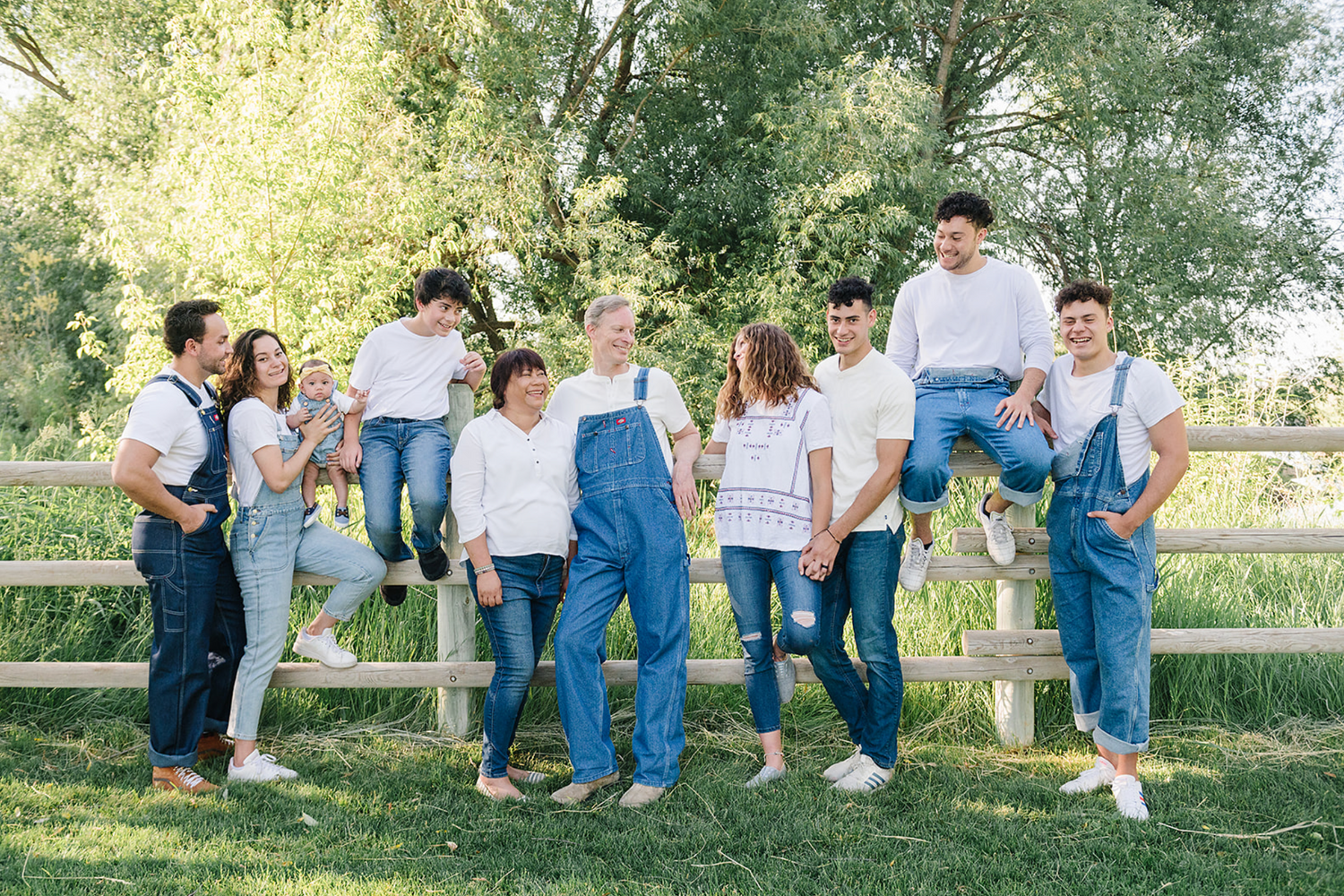Group in denim overalls and white shirts gathered casually by a wooden fence in a sunny outdoor setting.
