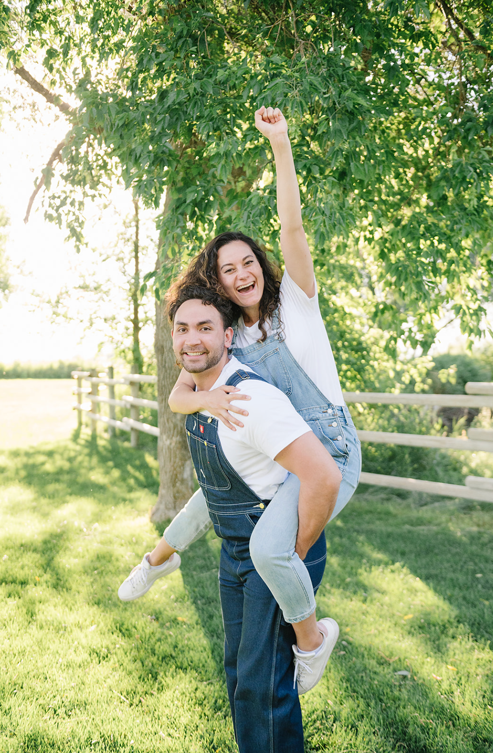 A playful couple enjoying a sunny day outdoors, with piggyback ride and laughter amid green trees and a wooden fence.