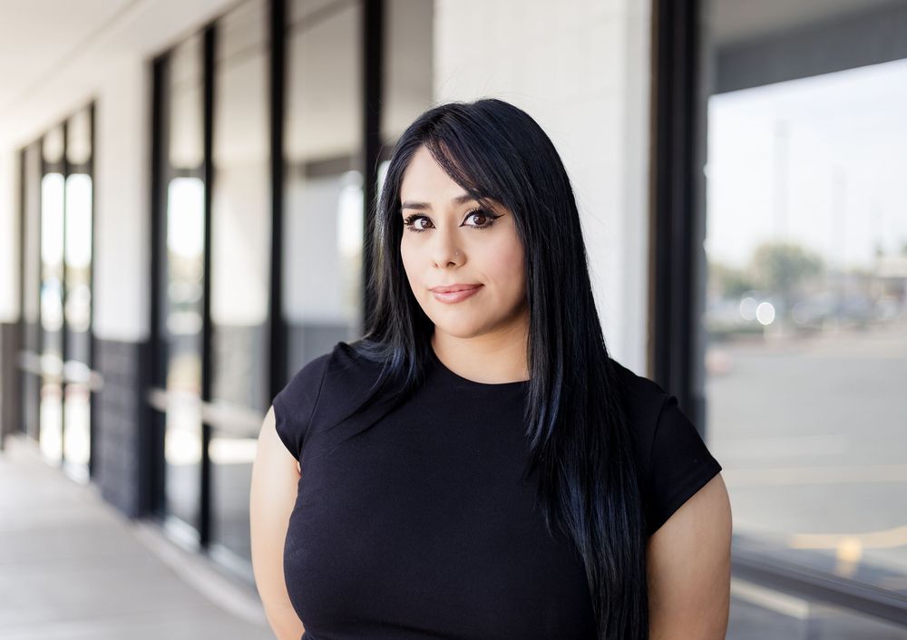 Person with long straight black hair wearing a black top poses thoughtfully against a storefront window.