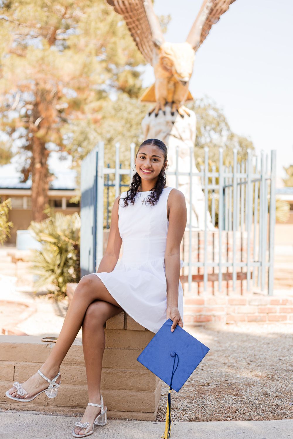 Graduate in white dress sitting on brick ledge holding blue diploma cover on sunny outdoor campus.