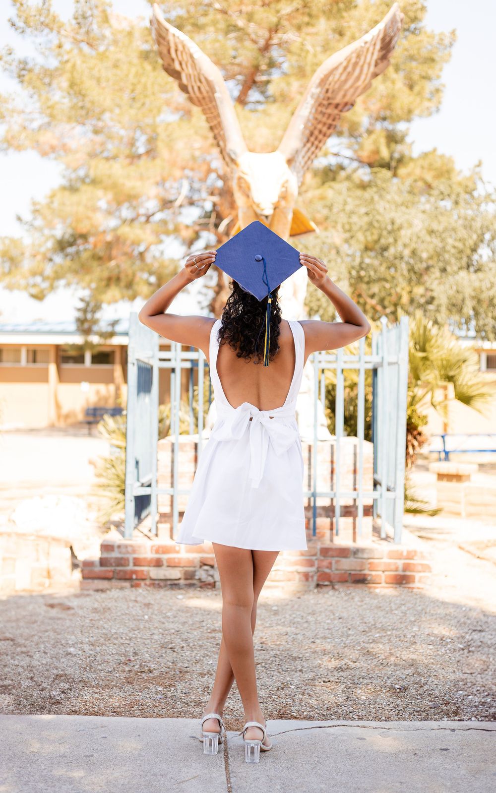 Graduate in white dress and cap holds arms up towards golden bird decoration in sunny outdoor setting.