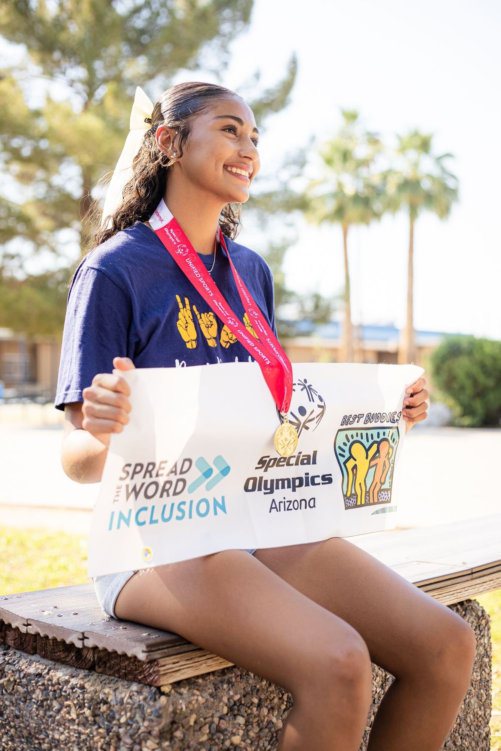 A cheerful participant wearing a navy Special Olympics shirt and medal sits outdoors holding a 'Spread Word Inclusion' banner.