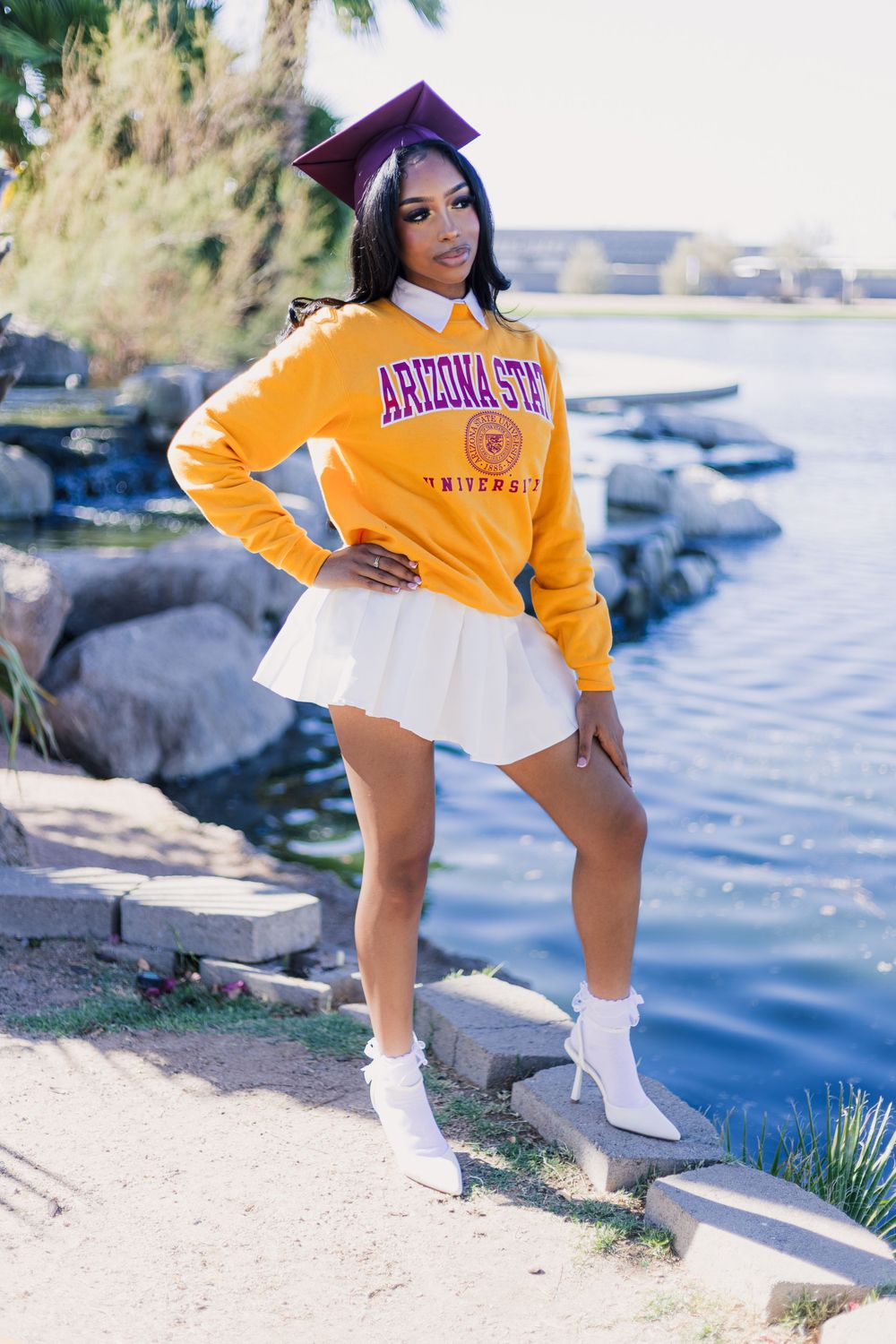 Graduate in yellow Arizona State University sweatshirt and white skirt poses near water with graduation cap.