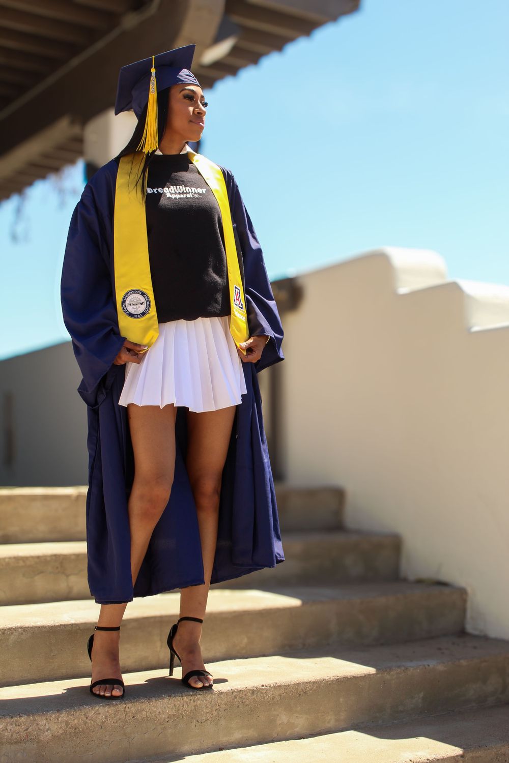 Graduate poses on stairs wearing navy cap and gown with yellow stole and white skirt.