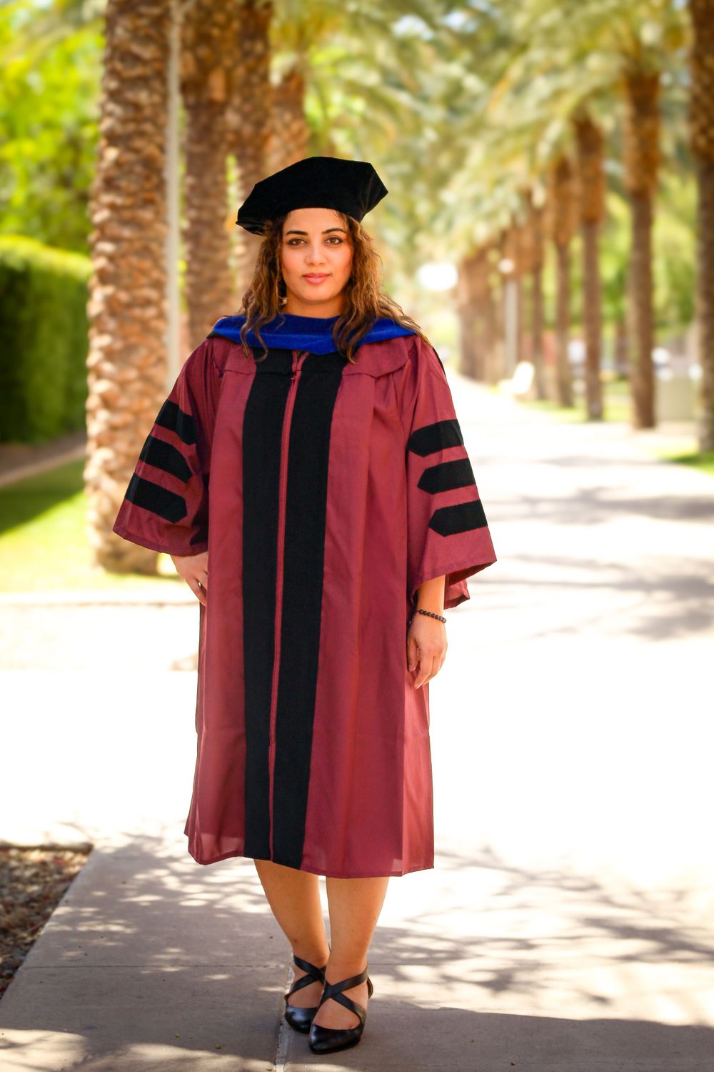Graduate in burgundy doctoral regalia stands on palm tree-lined pathway.