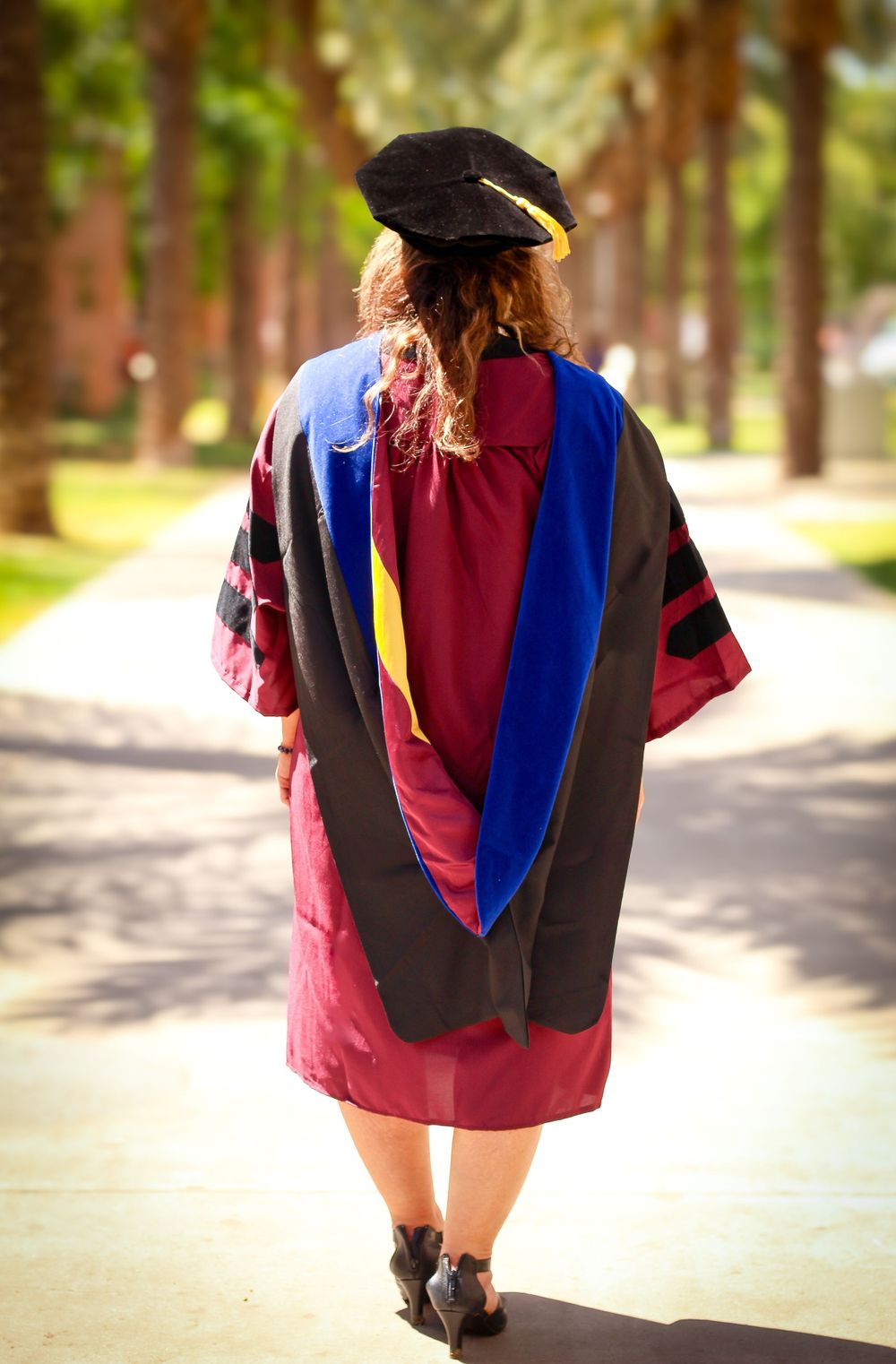 Graduate in red academic regalia and doctoral hood walking away on tree-lined path.