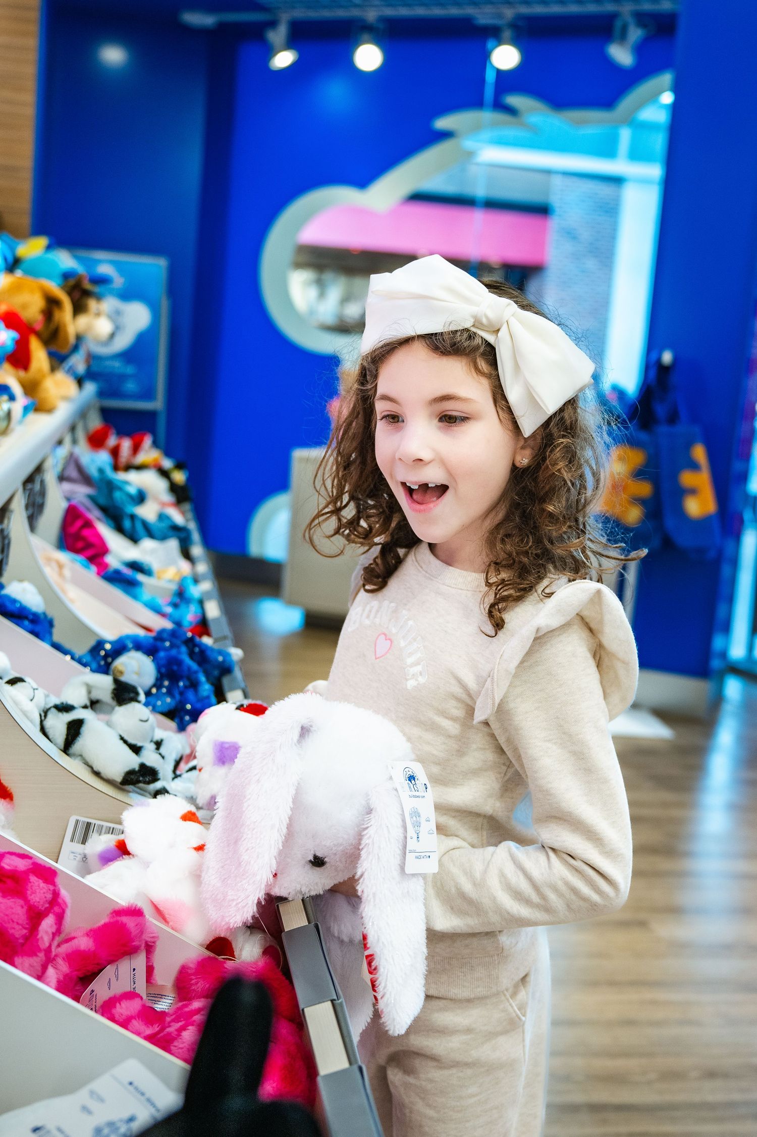 A young shopper wearing a white hair bow and cream sweater looks excited while holding a plush toy in a retail store.