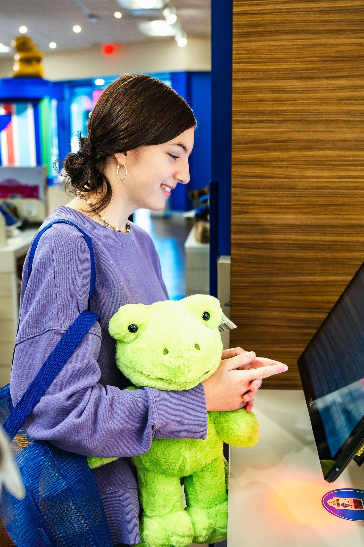 Person in purple sweater smiling while holding a bright green plush frog toy in a retail setting.