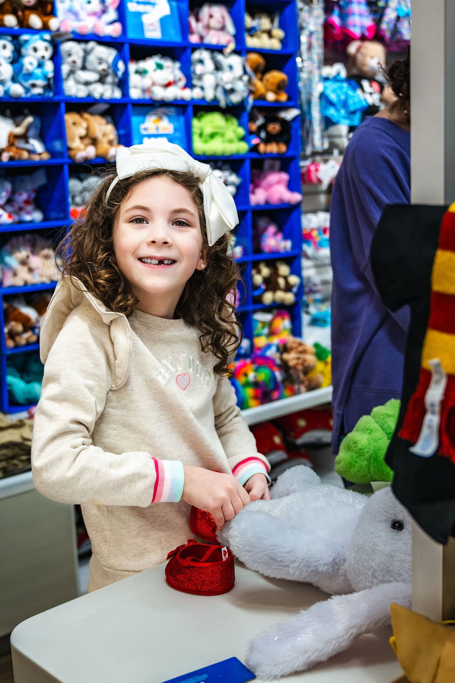 A child smiles while playing at a stuffed animal store with colorful plush toys displayed on shelves in the background.