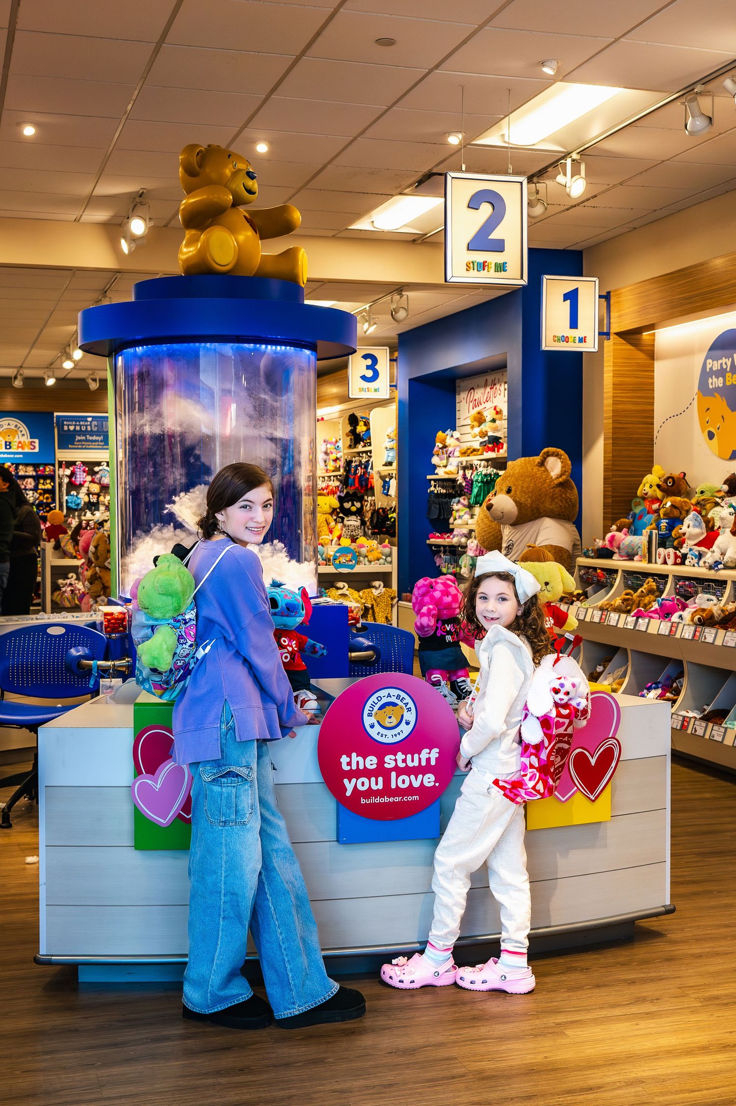 People browse inside a Build-A-Bear Workshop retail store with numbered stations and colorful plush toys on display.