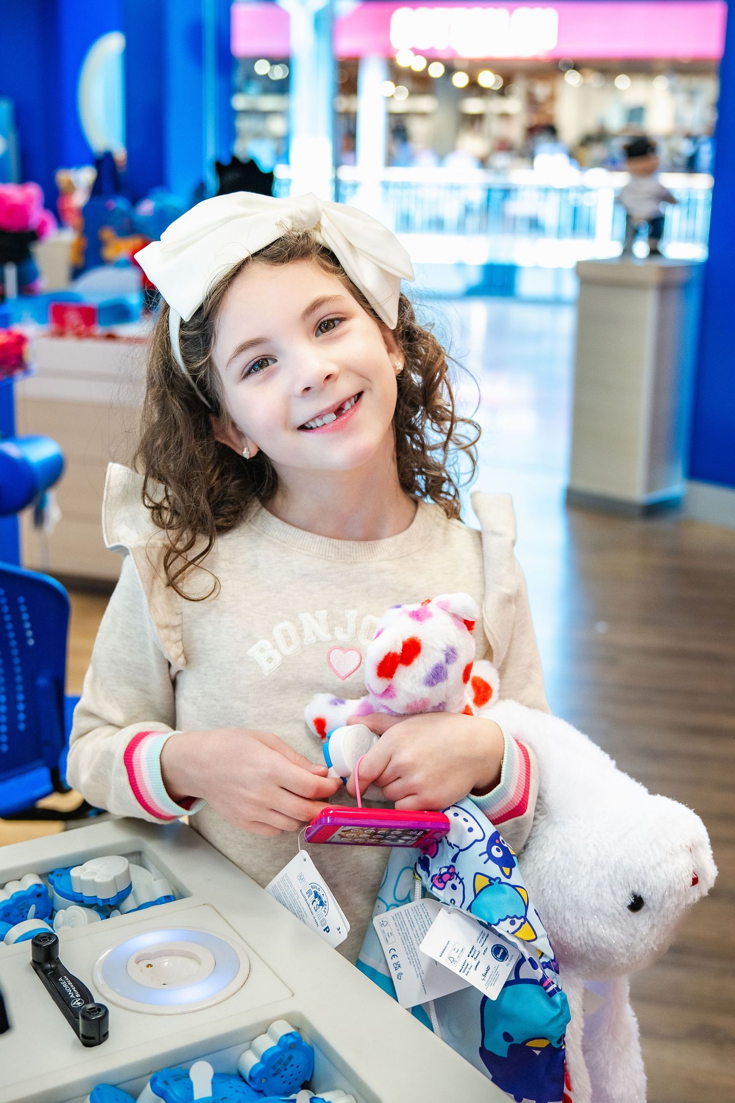 At Build-A-Bear Workshop, a joyful child wearing a white bow and gray sweater holds stuffed animals at a workstation.