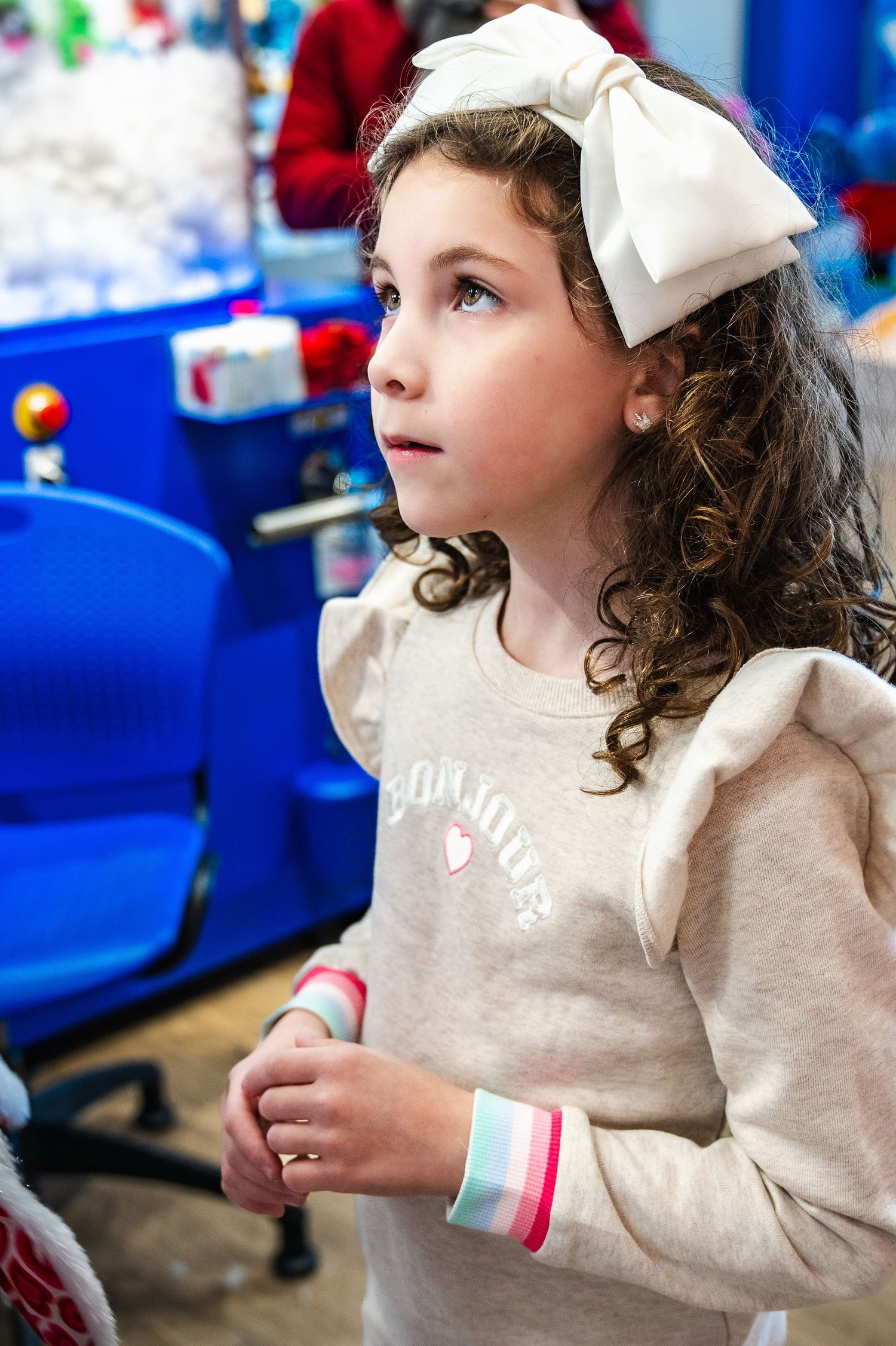 Young person with curly hair wearing a light beige sweatshirt and white hair bow looking to the side.