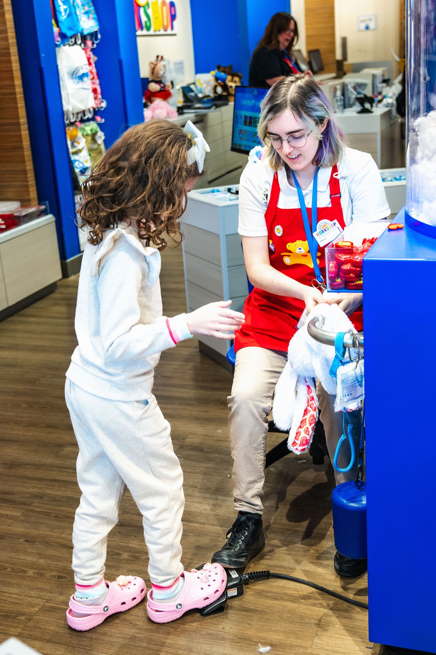 At a Build-A-Bear Workshop, a customer in white interacts with a store employee wearing a red apron on the shop floor.