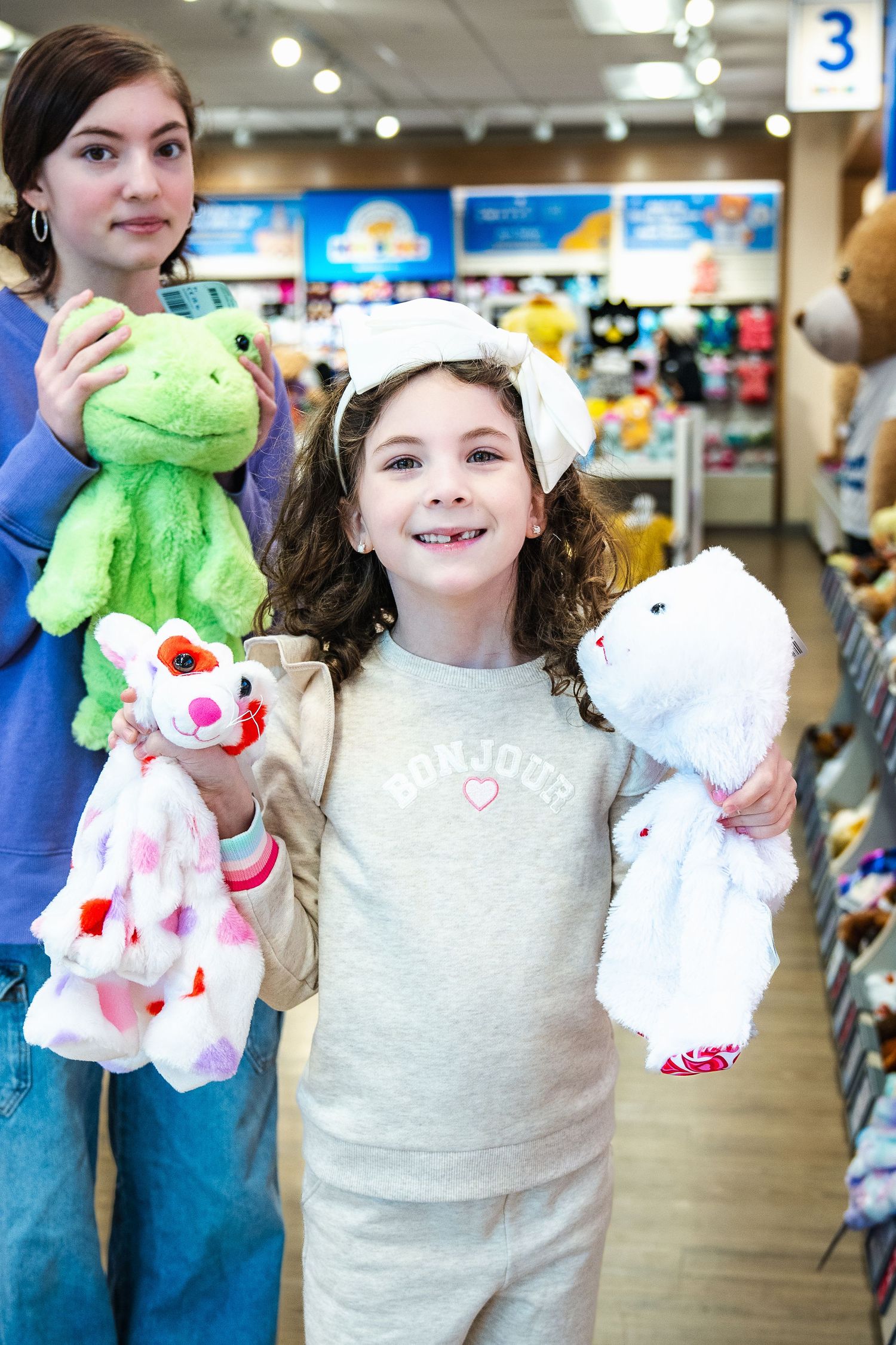 A parent and child hold colorful plush toys including a green frog and white bears while shopping in a toy store.