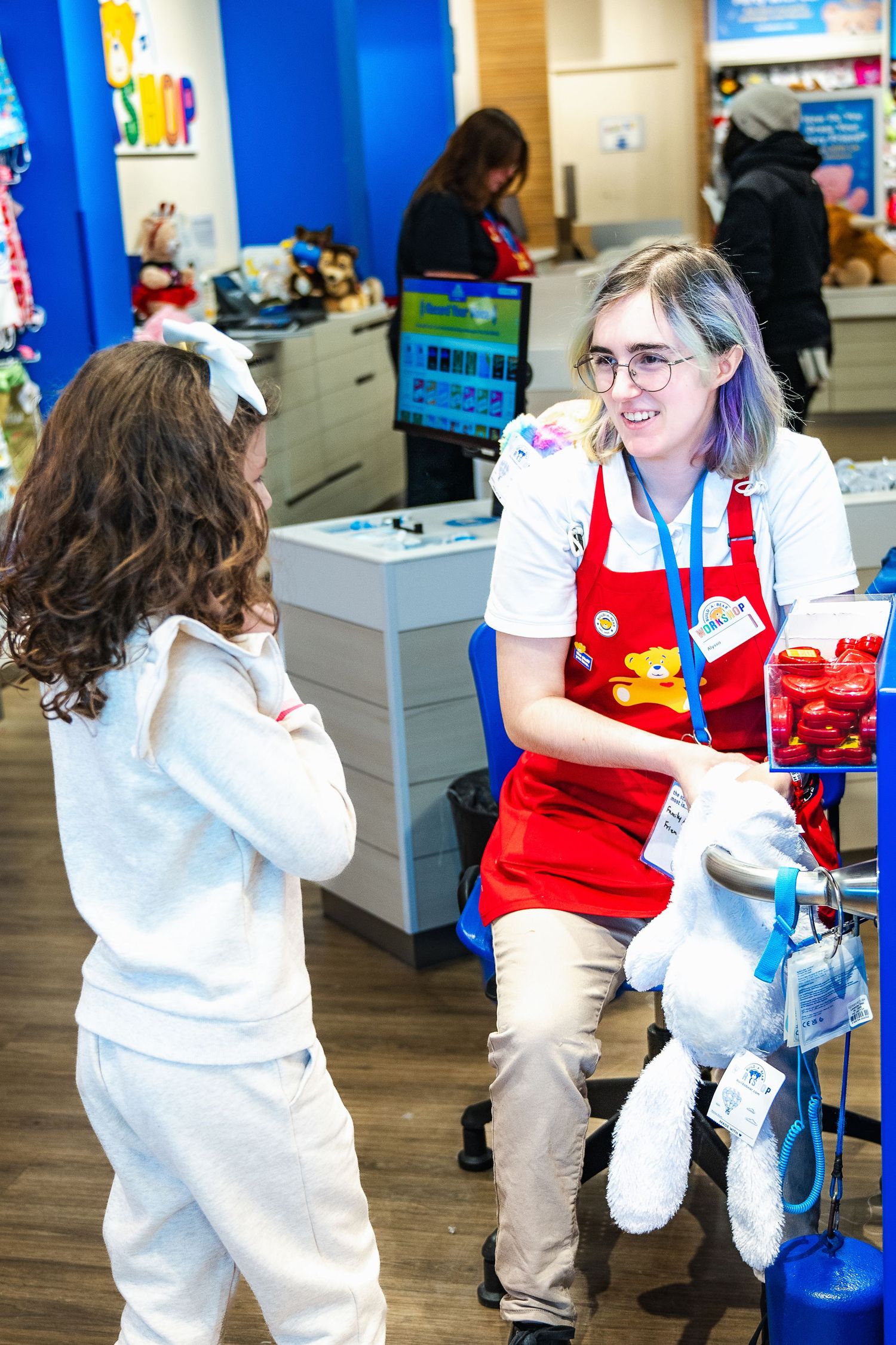 Store employee in red overalls and nametag interacting with a customer in a retail setting with blue walls and displays.