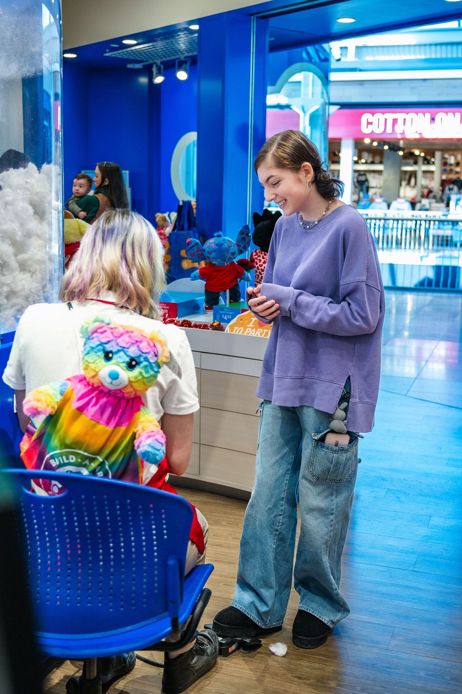 Customers stand at a counter in a blue-lit shopping mall with a vibrant tie-dye shirt visible in the foreground.