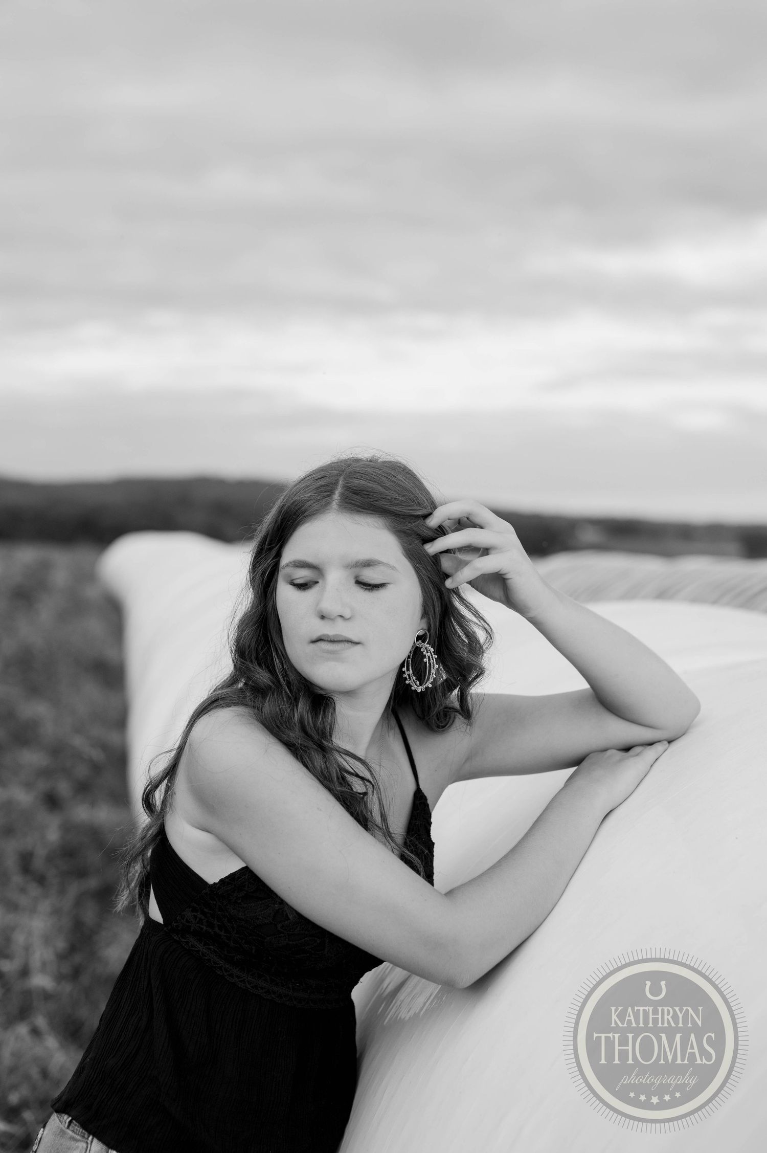 Black and white portrait leaning against round bales with eyes closed in dramatic lighting.