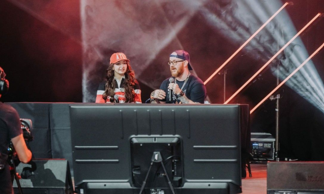 Two people sit at a desk at an event with dramatic red lighting and white spotlights beaming in the background.