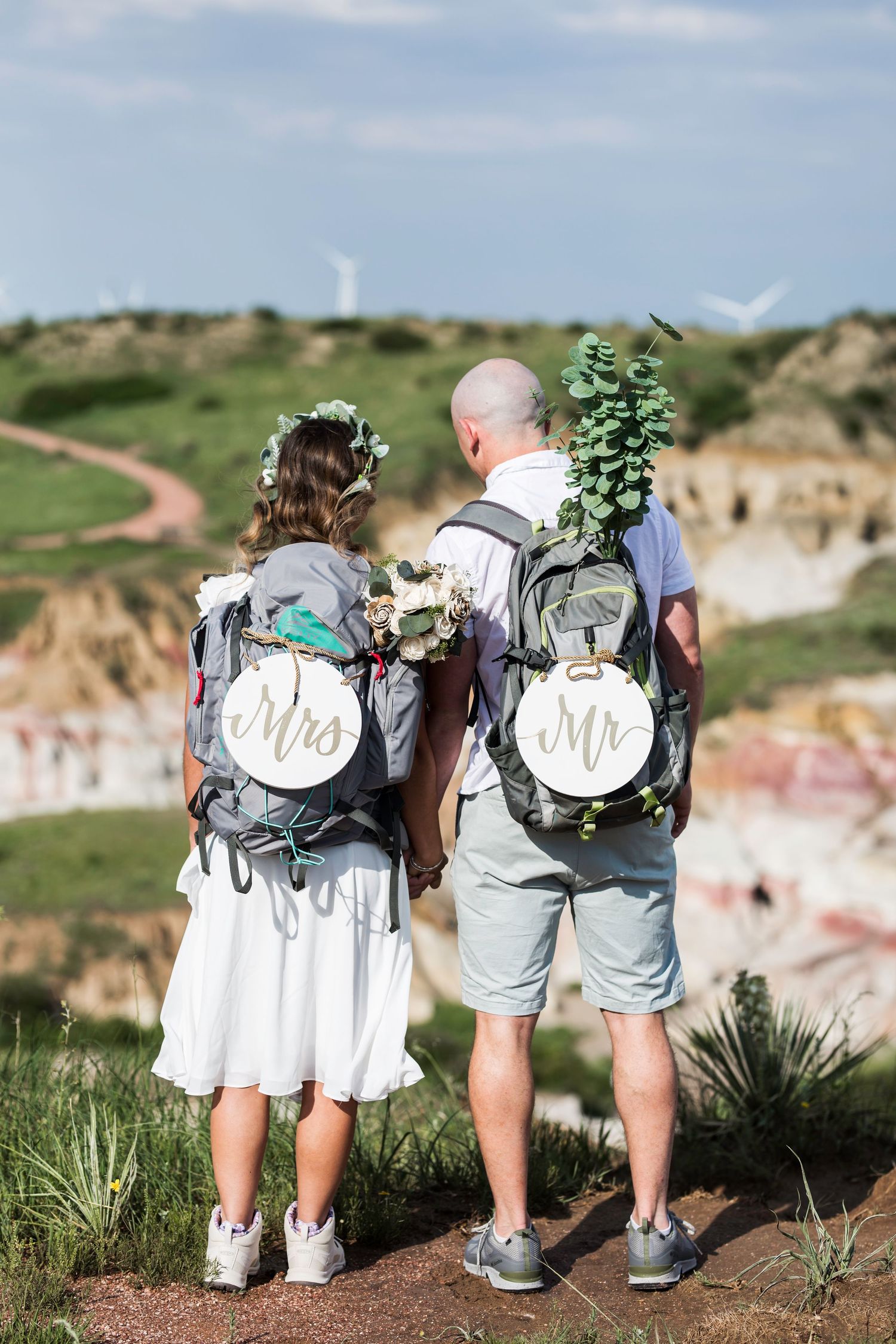 Couple wearing matching Mr and Mrs backpack signs standing together on hilltop overlooking scenic landscape.
