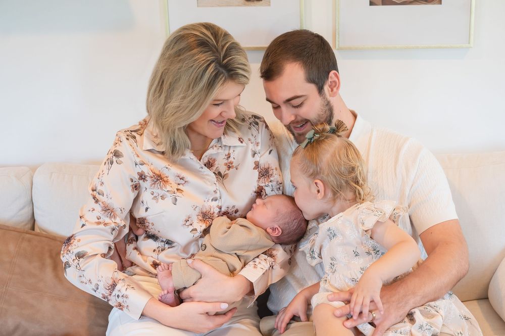 A happy family of four sits on a couch, parents smiling at their newborn baby while a toddler looks on.