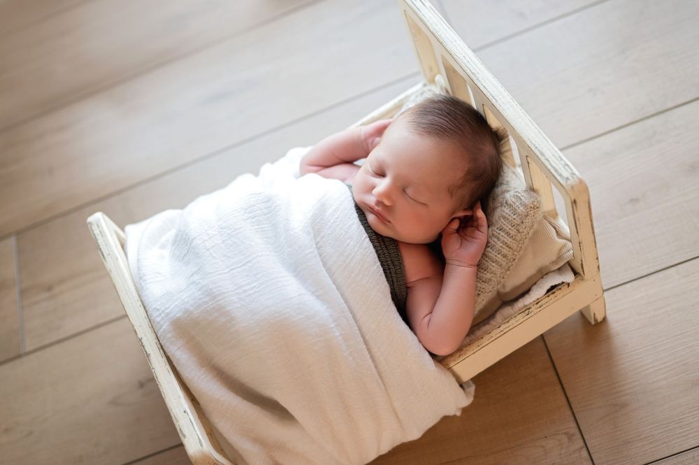 Peaceful newborn baby sleeping wrapped in white knit blanket in a small wooden prop bed on hardwood floor.
