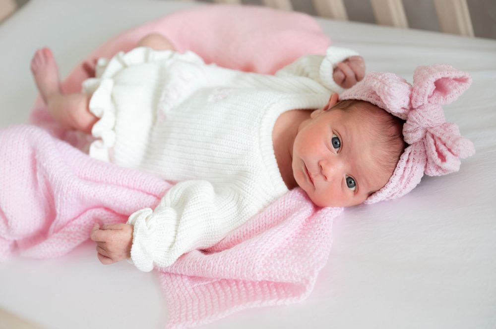 Newborn baby in white outfit lying on pink blanket with matching pink bow headband accessory.