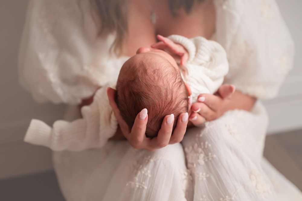 Hands gently cradle a newborn baby wrapped in soft white fabric against a light background.