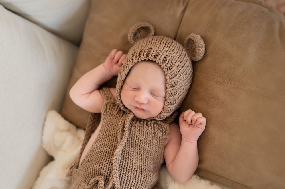 Sleeping newborn baby wearing a knitted brown bear bonnet and romper outfit, resting on a cozy beige sofa.