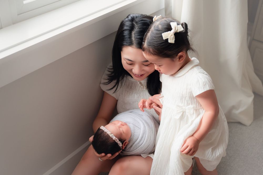 A mother sits by a window holding her newborn while her young daughter in a white dress looks on adoringly.