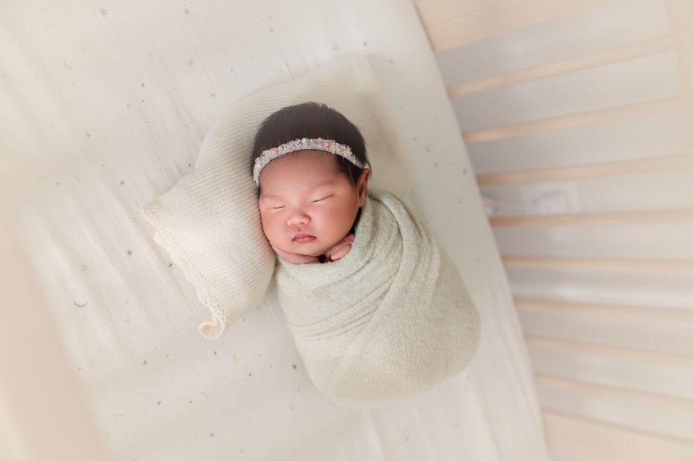 Newborn baby swaddled in soft mint wrap with brown headband, sleeping peacefully on white blanket.