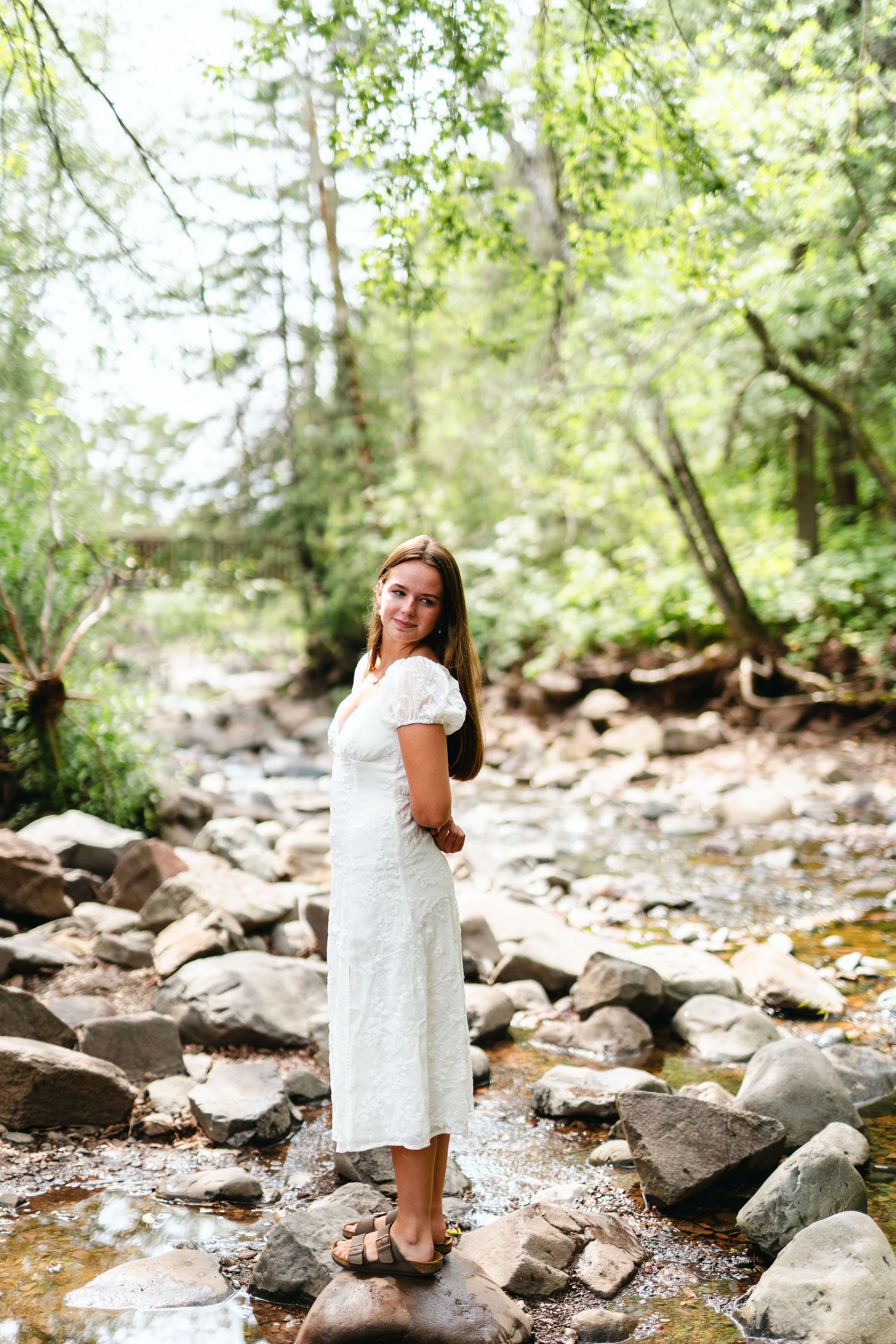 Person in white dress stands on rocky creek bed surrounded by lush green forest on sunny summer day.