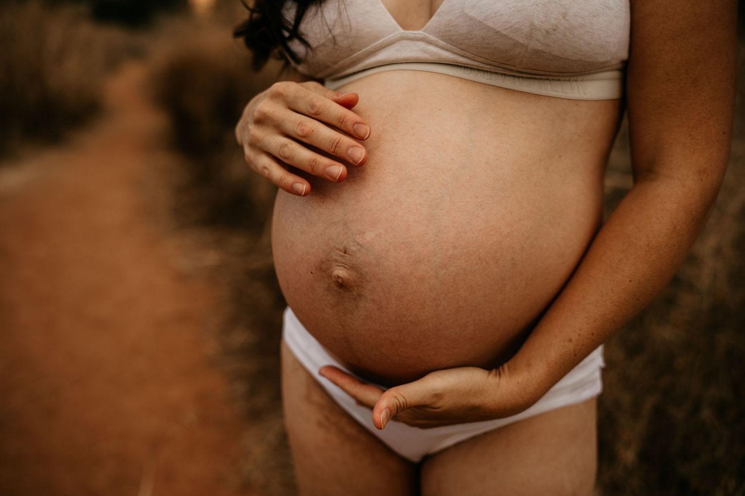 Pregnant woman cradling her bare baby bump outdoors on a natural earthy path.