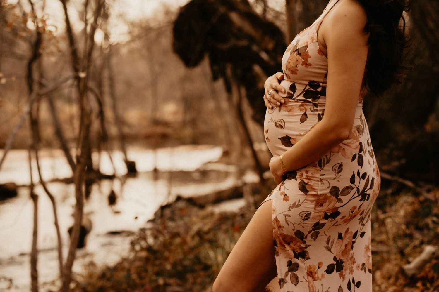 A maternity photo shoot in a floral dress by a waterside in sepia tones.