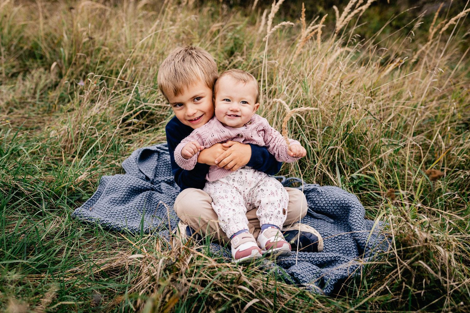 Zwei Kinder sitzen während eines Fotoshootings im Freien zusammen auf einer blauen Decke im hohen Gras.