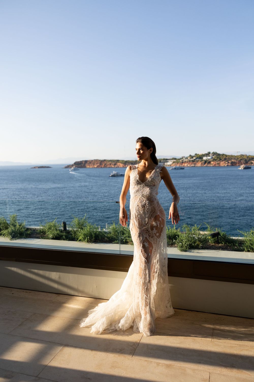 Elegant lace wedding gown silhouetted against scenic ocean coastline view from luxury balcony terrace.