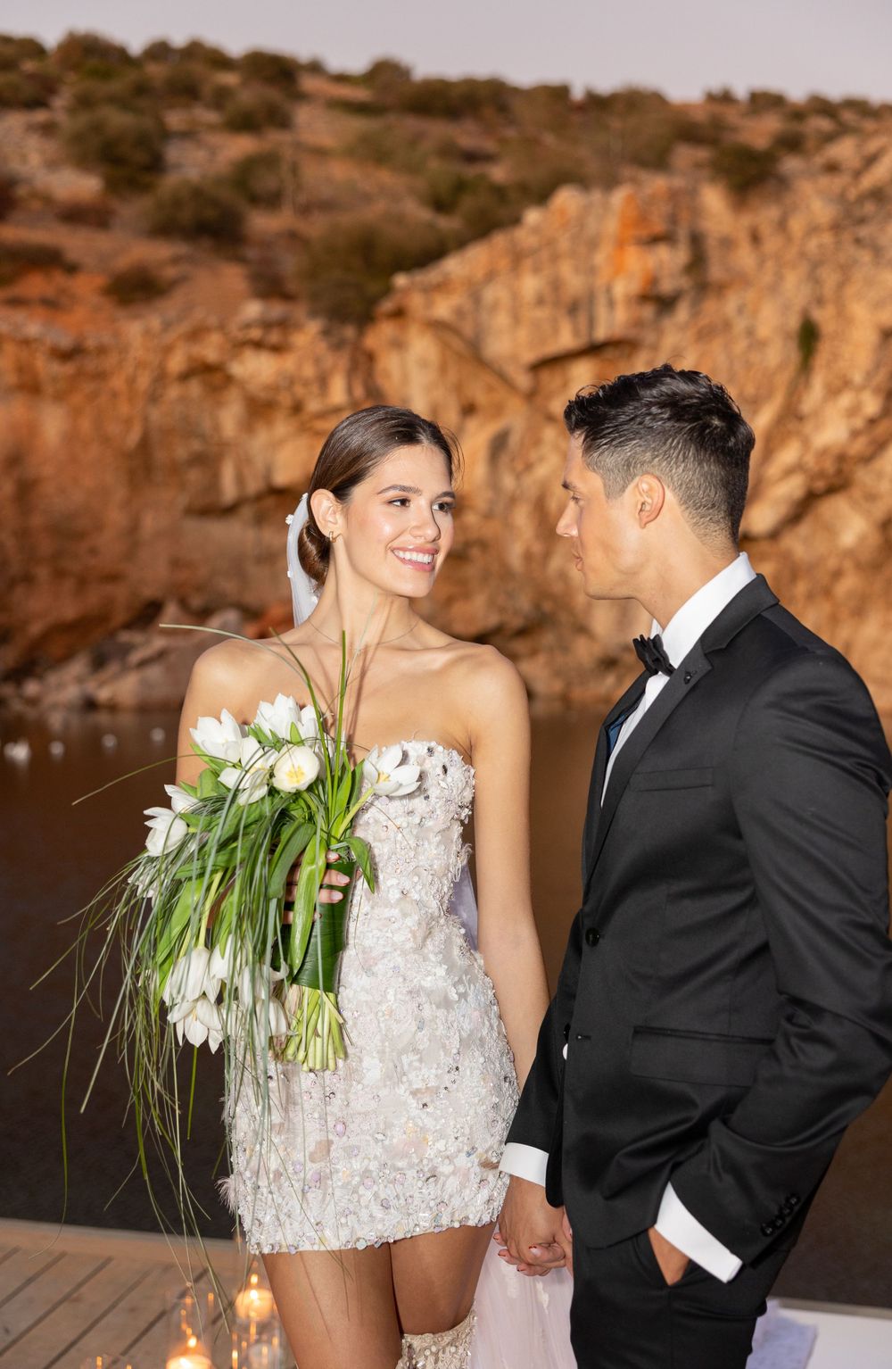 A couple in formal attire stands on a deck with candles and rocky cliffs in background.